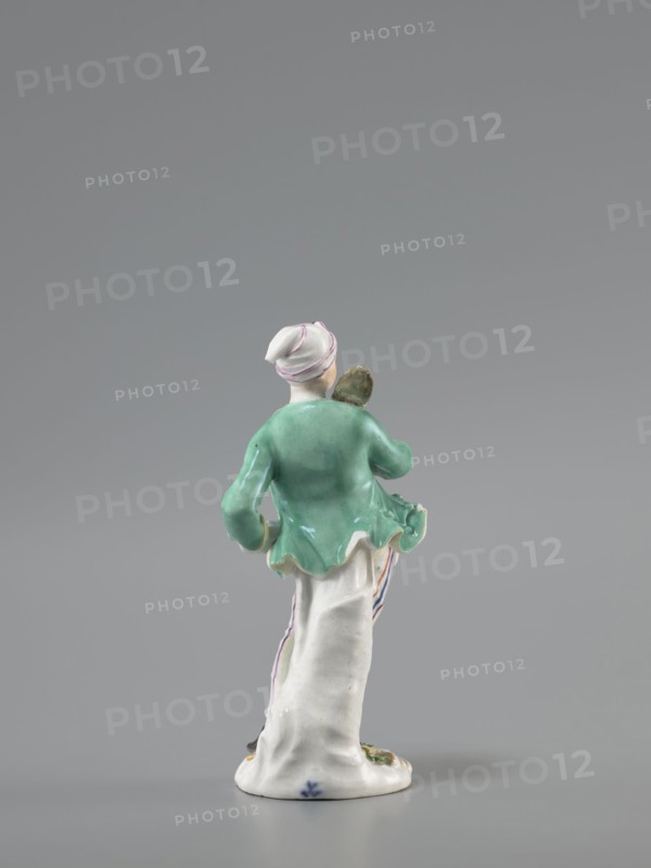 Man with a Potato Masher, Reinicke, Peter 1715–1768 modeler?; Koenigliche Porzellain Fabrique, Meissen 1710–1763 manufacturer. On a flat, circular pedestal with rocaille merging into a tree trunk stands a young man leaning on the trunk at waist level. In contrapposto, head turned to the right, he holds a short oval board with a handle—an instrument for stewing potatoes—resting on his right shoulder. Dressed in trousers with colorful thin stripes tied loosely at the waist with a belt, black shoes with a buckle, a short green jacket with box pleats, a pink waistcoat with gold braiding, and a soft cap similar to a nightcap. Ca. 1755, Saxony; Meissen. Figurine; height 13.5 cm., A small porcelain figurine shown from the back on a gray background, featuring a figure wearing a white head covering with thin pink stripes, a green jacket with a flared hem, and a long white skirt with subtle colored stripes at the side; the figure holds a small brownish object near the shoulder and stands on a wh...