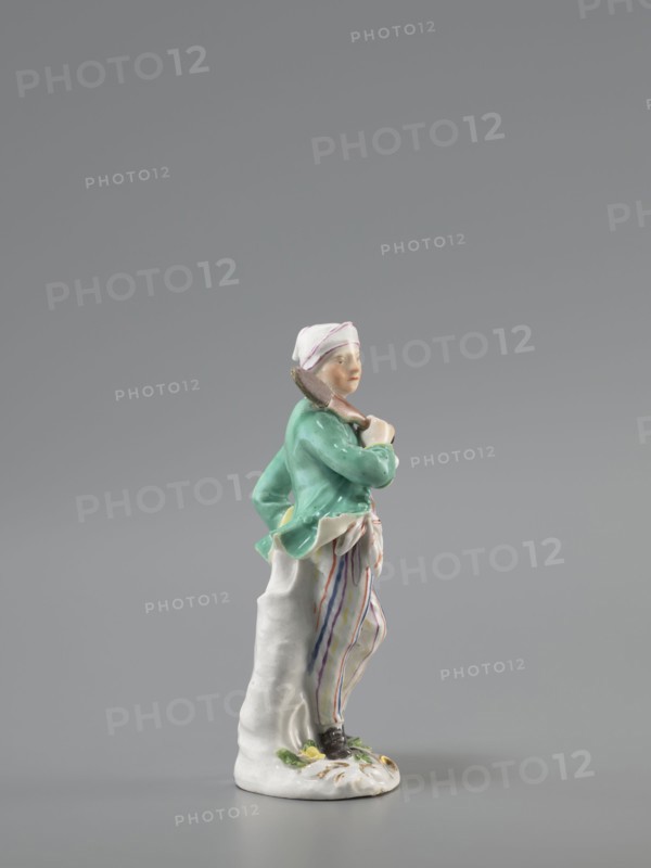 Man with a Potato Masher, Reinicke, Peter 1715–1768 modeler?, Koenigliche Porzellain Fabrique, Meissen 1710–1763 manufacturer, figurine. On a flat, circular pedestal with rocaille merging into a tree trunk stands a young man leaning on the trunk at waist level. In contrapposto, head turned to the right, he holds a short oval board with a handle—an instrument for stewing potatoes—resting on his right shoulder. Dressed in trousers with colorful thin stripes tied loosely at the waist with a belt, black shoes with a buckle, a short green jacket with box pleats, a pink waistcoat with gold braiding, and a soft cap similar to a nightcap. Ca. 1755; Meissen, Saxony. Figurine, height 13.5 cm., A small porcelain figurine of a standing person wearing a white head covering with a faint pink stripe, a green jacket, and baggy white trousers with vertical stripes in purple, orange, and blue, holding a brown object over one shoulder and leaning against a white textured support, standing on a white base...