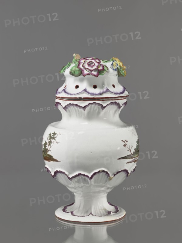 Pot-pourri vase with lid by Johann Georg Buchwald Cologne; 1769–1771. Porcelain vessel with a hemispherical body slightly narrowing at the top, almost flat shoulders, and a wide obliquely sloping neck; low stem widening into a round, wide foot. Neck, base of the belly, and foot are bordered by a wreath of applied shell ornament painted purple. Two painted landscapes with buildings amid trees on the body. Hat-like cover with brim enclosed in shell ornament; top features applied colorful flowers with a floral handle. Subject: flowers, pot-pourri, landscapes, architecture. Date: 1769–1771. Place: Cologne, Germany. Height 37 cm; width 26 cm., A white ceramic vase with a scalloped form and a pedestal base, decorated with painted accents in purple and brown and small painted landscape motifs in green and brown on its body, topped with a removable lid adorned with molded and painted flowers and leaves in purple, yellow, green, and blue.