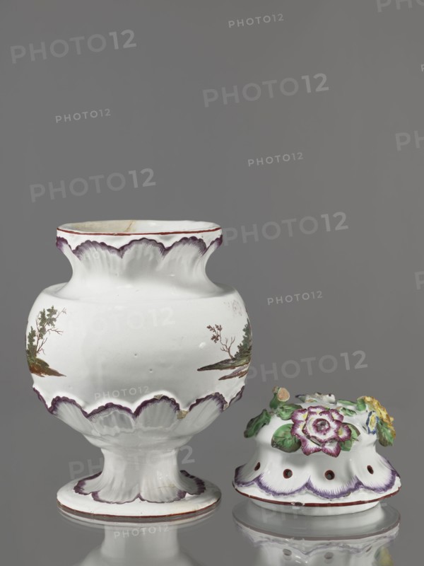 Pot-pourri vase with lid by Johann Georg Buchwald Cologne; 1769–1771. Hemispherical body slightly closing at the top, almost flat shoulders, and a wide neck that slopes obliquely. Low stem widening into a round, wide foot. Neck, base of the belly, and foot enclosed in a wreath of plastic shell ornament painted in purple. Two landscapes with buildings against a background of trees on the body. Hat-like cover with a brim enclosed in a shell ornament; top with a plastic decoration of colorful flowers and a floral handle. Materials: porcelain pot-pourri vessel. Date: 1769–1771. Place: Cologne, Germany. Height 37 cm; width 26 cm., A white ceramic vessel with a flared scalloped rim and pedestal base sits next to a separate domed lid decorated with molded flowers; the vessel has painted landscape scenes in green and brown on its rounded body and purple edging along the rim, scallops, and base, while the lid features molded flowers and leaves in green, purple, yellow, and pale blue with purple...