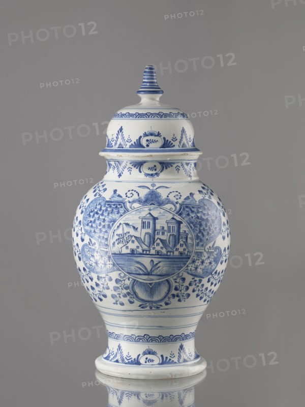 Vase with lid, Fayence-Manufaktur Muenden faience factory; 1732–1854 Hannoversch Münden, architecture, Méyet, Leopold 1850–1912; birds, plant motifs. Baluster vase with a short, slightly flared neck, pear-shaped belly, on a conical foot, with a domed lid terminating in a conical handle. Around the lid, neck, and foot is a pattern of triple semicircles, fir-tree and oval plant motifs and borders. On the front, in a circular cartouche, an architectural view with two towers and three houses with gable roofs. The cartouche is topped with a plant palmette with a circular motif at the bottom and richly ornamented on both sides with a scale mosaic, Chinese latticework, double volutes, and two birds at the top. Branches with flowers, fruits or oak leaves radiate horizontally from the cartouche. On the back are small stars and a bipartite shoot motif. 3rd quarter of the 18th century, Germany; Hannoversch Münden. Vessel; height 41.8 cm, width 21.0 cm., A lidded ceramic jar with a bulbous body an...