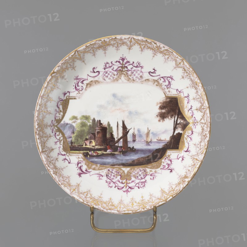 Jug plate, Koenigliche Porzellain Fabrique, Meissen circa 1720–1735, Meissen, Saxony. Porcelain shallow bowl plate in an oval, concave-convex, multi-part reserve in a mirror, bordered with gilded lace and supplemented with purple leafy volutes with fields of Chinese lattice. Central colorful landscape miniature with staffage: a bay with a sailing boat, architecture including a round building on the shore, and two sailing boats in the distance. Around the rim a trail of gilded, serrated lace. On the reverse of the collar three Indianische Blumen branches. Height 3.8 cm; diameter 18 cm. Maker attribution: Koenigliche Porzellain Fabrique, Meissen 1710–1763., A round ceramic plate with a central painted scene framed by ornate decorative borders; the scene shows a waterfront with sailing boats and a small stone building near trees, and a shoreline with a tree on one side, painted in muted tones of brown, green, blue, and gray with touches of reddish-brown; the plate rim is decorated with in...