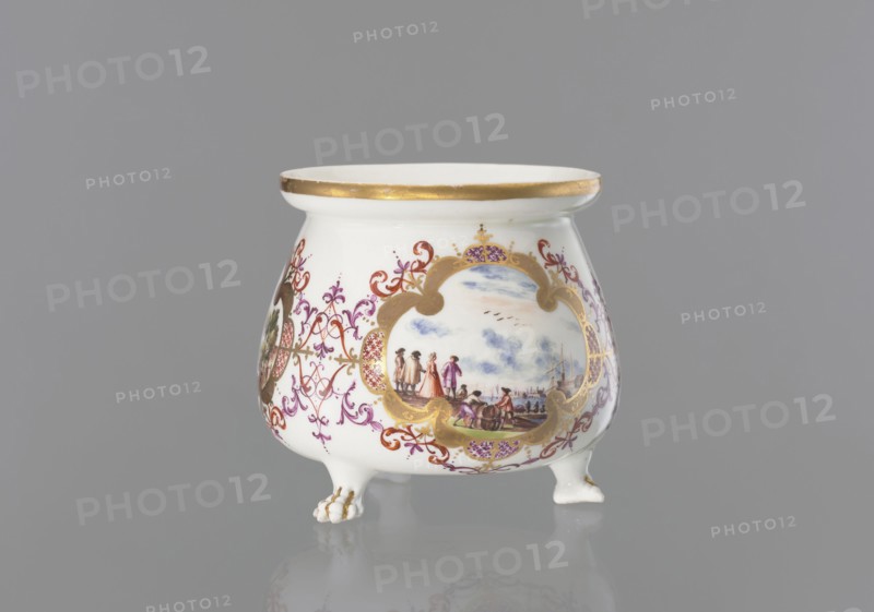 Lion's paw pot with miniatures, Koenigliche Porzellain Fabrique, Meissen ca. 1730, maker: Meissen 1710–1763. Whipped cream pot vessel with squat belly, wide collar around the rim; volute, segmental handle gilded along the spine; on three legs shaped as lion's paws. Body decorated with three oval/quadrangular reserves surrounded by gilded frames with ovals of colorful Chinese latticework and purple-brick-red leaf motifs and Regency volutes—each reserve contains a colorful landscape miniature by the water with staffage: 1 a barrel-wheeler and four men at the quay, 2 two men with cargo at the shore and a woman and three men standing nearby, 3 four men waiting with luggage and sailing boats at the shore. Between the miniatures are two brick-purple Regency compositions with gilding; lion's paws and outer edge of the collar are gilded. Origin: Meissen, Saxony, Germany., A small white porcelain vessel with a rounded body and flared rim sits on three short feet, each foot shaped with a paw-lik...