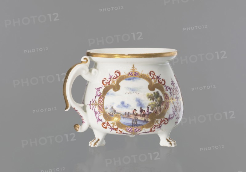Lion's paw cream pot with miniatures, Meissen porcelain factory, ca. 1730. Porcelain whipped-cream pot with squat belly and wide collar; volute, segmental handle gilded along the spine; on three legs shaped as lion’s paws. Body decorated with three oval/quadrangular reserves surrounded by a gilded frame with ovals of colorful Chinese latticework and purple-brick-red leaf motifs and Regency volutes—each reserve with colorful landscape miniatures by the water with staffage: 1 a barrel-wheeler and four men at the quay; 2 two men with cargo at the shore and a woman and three men standing nearby; 3 four men waiting with luggage and sailing boats at the shore. Between the miniatures, two brick-purple Regency compositions with gilding; lion’s paws and outer edge of the collar gilded. Saxony, Meissen, Germany., A white porcelain cup with a curved handle and three small clawed feet, decorated with gold trim and gold accents on the handle and feet, features an ornate framed scene painted on its ...
