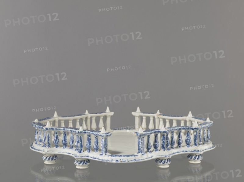 Base for a tabletop tray. Oval architectural tray serving as a base for a tabletop tray. Carved with 4 leaves, with a wavy edge — alternately concave and convex. Supported by 8 round, spirally fluted legs. Around the edge is a baluster gallery with balusters interwoven with small columns and pillars, topped with cones. Painted in a blue Regency arabesque. Inner walls and base white. 1st half of the 19th century; possibly 18th century France, Strasbourg . Vessel. Height 11.0 cm; width 33.0 cm; depth 45.0 cm., A decorative ceramic object with a pale white base and blue painted ornamentation sits on a reflective surface; it has a low, irregularly shaped platform surrounded by a balustrade of small turned posts and pointed finials, with the blue decoration forming floral and linear patterns along the rim, posts, and rounded feet, and the overall color scheme includes white and blue against a gray background.