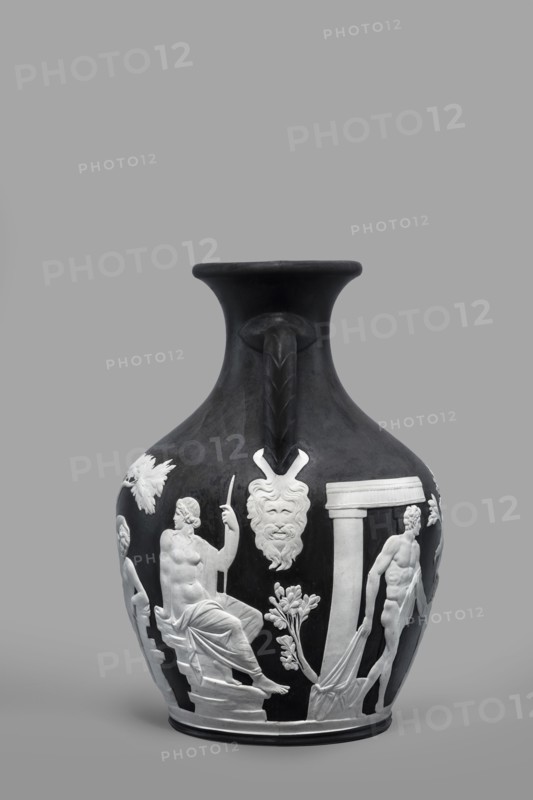 Portland Vase, copy, Josiah Wedgwood & Sons Stoke-on-Trent; pottery manufactory; 1790–1895. Maker: Wedgwood, Josiah 1730–1795. Pelike-shaped vase, black with white reliefs white rendered from a black background depicting: 1 Atia, mother of Emperor Augustus, Apollo and Neptune; 2 Hecuba wife of Priam with a torch in her hand, surrounded by Venus and Paris. At the bottom, Paris, lost in thought. 19th/20th century. Staffordshire, Stoke-on-Trent, England. Vase. Height 26.6 cm; width 19 cm., A dark vase with a flared rim and a single ornate handle rising from the neck sits against a plain gray background; the vase is primarily black with raised white reliefs depicting classical figures and architectural elements, including a seated draped figure holding a staff, a standing partially draped male figure, a stylized bearded mask hanging below the neck, floral motifs, and column-like forms, with contrasts of black and white tones throughout.