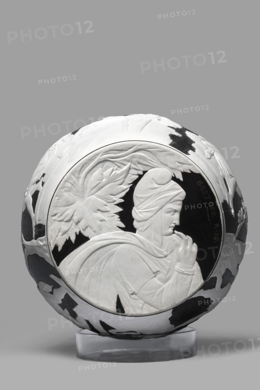 Portland Vase, copy, Josiah Wedgwood & Sons Stoke-on-Trent; pottery manufactory; 1790–1895. Pelike-shaped vase in black with white rendered from a black background reliefs depicting: 1 Atia, mother of Emperor Augustus, Apollo and Neptune; 2 Hecuba wife of Priam with a torch in her hand, surrounded by Venus and Paris. At the bottom, Paris, lost in thought. 19th/20th century, Staffordshire, Stoke-on-Trent, England. Vase; height 26.6 cm, width 19 cm., A round sculpted object with a carved profile of a person in relief on one face, shown against a plain gray background; the object is primarily white with several areas of black contrasting patches and details, the central medallion features the figure with draped garments and a head covering, surrounded by carved leaf-like shapes, and the piece rests on a translucent clear stand.