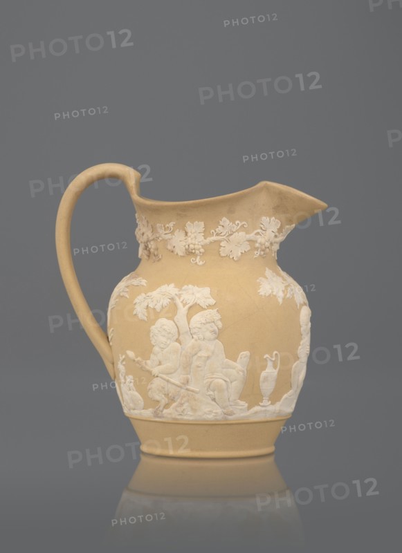 Oinochoe jug, cane ware pottery, beige-sand with a ribbon handle; around the belly, white reliefs: a group of three putti — two naked putti, tied with drapery, carry on a saddle a naked putto with a thyrsus and a vine; a naked putto with a panpipe is seated under a tree; a putto with two amphorae is seated under a tree; a satyr and a putto with two amphorae, a basket of vines; under the rim around the neck, a trail of vine shoots with fruit. Inside, a glazed jug. Maker: Wedgwood, Josiah 1730–1795; Etruria factory pottery manufactory. Date: c. 1815–1820. Place of manufacture: Stoke-on-Trent, Staffordshire, England. Subject: bacchanalia. Function: vessel. Height: 12 cm., A beige ceramic pitcher with a curved handle and a pointed spout, decorated with raised white reliefs depicting a group of cherubic figures, foliage, and a vase, set against a solid gray background; colors visible include beige, white, and gray.
