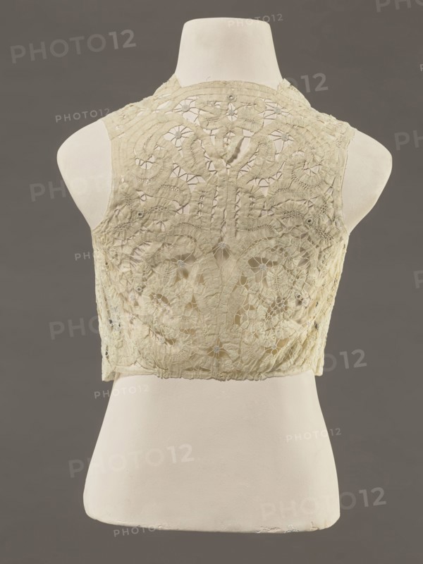 Bodice, lace, bodices, women's clothing, fashion. In a pattern of rosettes connected by large spiders and heads. Pattern of stylized floral motifs. 19th century, France. Clothing accessory. Height 36 cm, circumference 63 cm., A cream-colored lace sleeveless bodice displayed on a white mannequin torso against a dark gray background, showing an intricate floral and geometric lace pattern with openwork areas and raised embroidered motifs, scalloped edges at the shoulders and hem, subtle signs of wear and irregularities in the fabric, and visible texture contrasts between the delicate lace and the smooth mannequin surface; visible colors include cream, white, and dark gray.