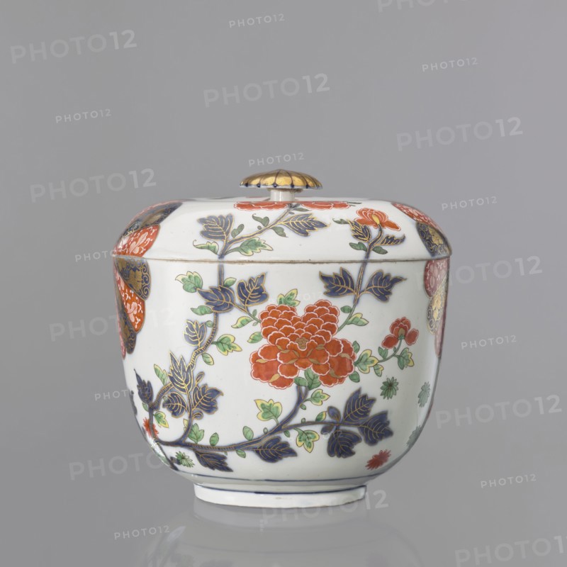 Vase with lid with Imari decoration, Kaiserliche Porzellanmanufaktur in Wien 1744–1864. Hemispherical vase with a separated bottom rim. Flat-arched lid with a button-rosette handle. Around the body, in a quadripartite arrangement, is Imari decoration continuing onto the lid: two fan-shaped 14-petal rosettes in brick red and cobalt blue with gilding, alternating with a fan-shaped cluster of lanceolate leaves with lotus flowers at the base of the bottom rim and two smaller tufts of leaves on the sides, and a peony shoot-branch with two full brick-red flowers and two buds; between the four motifs at the bottom are green and brick-red star-flowers and small leaves. Gilded rosette handle with a cobalt disc and a brick-red volute around the base. At the bottom, a double cobalt border with an inscribed signature in the center. Mid-19th century. 18th century, Austria, Vienna. Decorative vessel; height 20.5 cm, diameter 21.5 cm., A lidded ceramic vessel sits against a neutral gray background, s...