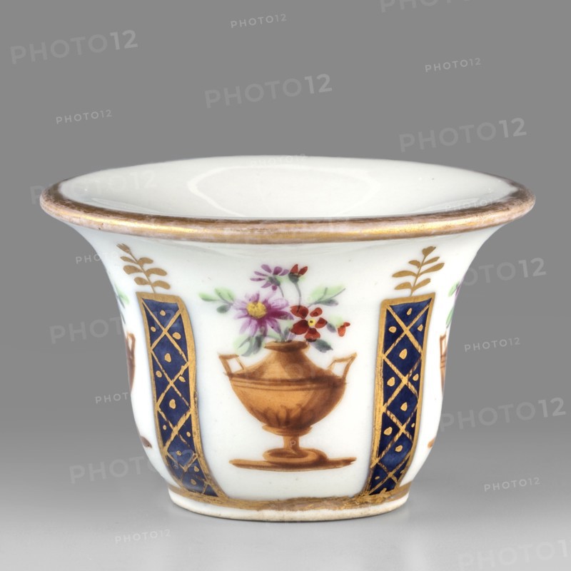 Toilet vessel for collecting tears or rubbing rouge?, Kaiserliche Porzellanmanufaktur in Wien 1744–1864. Bell-shaped porcelain vessel with a shallow circular recess; colorful decoration featuring four cobalt columns with gilded latticework alternating with four brown amphorae-vases containing floral arrangements; upper edge with gold borders. Identified as a vessel for collecting tears or rubbing rouge?. 1807, Vienna, Austria. Toilet accessory. Height 3.5 cm; width 5.5 cm. Maker: Kaiserliche Porzellanmanufaktur in Wien., A small white porcelain cup with a flared rim edged in gold shows painted decorations around its exterior: a stylized brown urn holding pink, purple, red, and yellow flowers with green leaves sits between vertical dark blue panels decorated with gold lattice and dot motifs, and small gold leafy sprigs appear above the panels; the cup rests on a short foot with a gold band near the base and the visible colors include white, gold, brown, dark blue, pink, purple, red, yel...