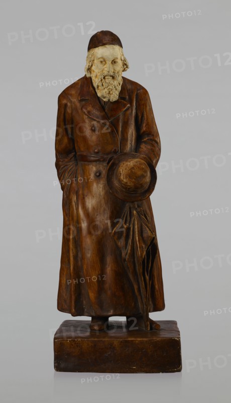 Figurine of a Jew, First Galician Factory of Artistic Faience and Terracotta Pacyków; 1912–1939. Figure of a Jew with sidelocks in a Crimean cap, dressed in a brown gabardine, holding a hat and umbrella in his left hand. Figure placed on a low plinth. 1st quarter of the 20th century, Pacyków. Figurine; height 33.7 cm, width 13.0 cm, depth 8.0 cm., A carved standing figure wearing a long coat and a cap, holding a round hat and a folded umbrella in front, with a detailed beard and hair; the sculpture rests on a rectangular base. The visible colors include shades of brown on the coat, hat, umbrella, base, and cap, a lighter beige on the face and beard, and a neutral gray background.