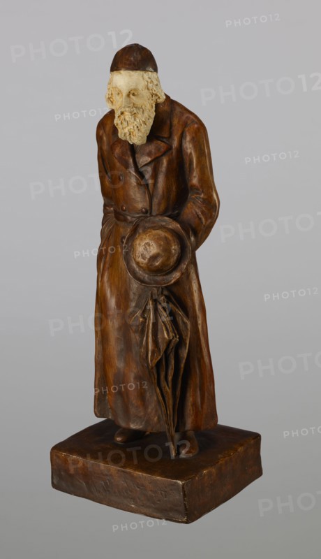 Figurine of a Jew, First Galician Factory of Artistic Faience and Terracotta Pacyków; 1912–1939. Figurine of a Jew with sidelocks in a Crimean cap, dressed in a brown gabardine, holding a hat and umbrella in his left hand. Figure placed on a low plinth. 1st quarter of the 20th century, Pacyków. Terracotta/faience figurine; height 33.7 cm, width 13.0 cm, depth 8.0 cm., A standing sculpted figure with a textured light beige head and beard and a darker brown cap, wearing a long brown coat that reaches near the base; the figure holds a round hat in front and an umbrella or folded cloth hanging down, all rendered in warm brown tones with variations of darker and lighter brown; the base is a rectangular brown plinth; the background is a flat light gray.