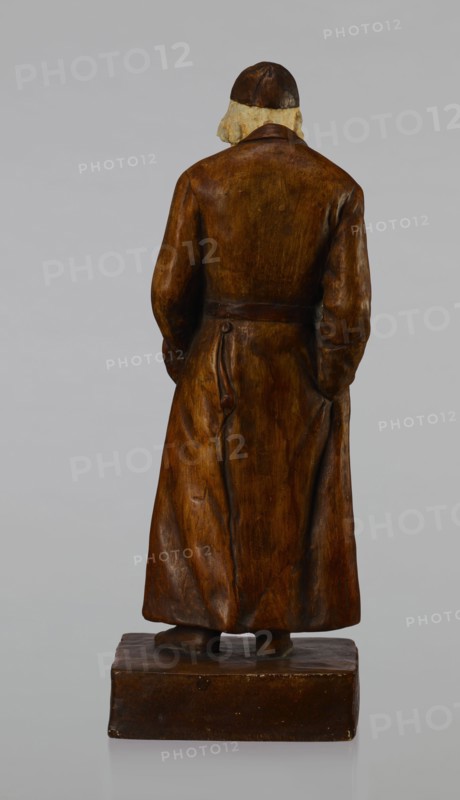 Figurine of a Jew, First Galician Factory of Artistic Faience and Terracotta Pacyków; 1912–1939. Figurine of a Jew with sidelocks in a Crimean cap, dressed in a brown gabardine, holding a hat and umbrella in his left hand. Figure placed on a low plinth. 1st quarter of the 20th century, Pacyków. Figurine; height 33.7 cm, width 13.0 cm, depth 8.0 cm., A carved figure seen from behind standing on a rectangular base, wearing a long coat and a close-fitting cap, with hair visible beneath the cap; the figure and base are rendered in brown tones with lighter beige for the hair, and the background is a uniform pale gray.