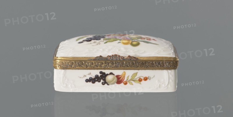 Snuffbox tin, Koenigliche Porzellan Manufaktur KPM; Berlin; 1763-1918 manufacturer, flowers, fruit, snuffboxes, miniatures. Snuffbox in the form of a rectangular box with a slightly arched lid, framed in gilded bronze, with a plant ornament around the outer edge of the lid and a lock, on hinges. Around the edge of the lid and the side walls of the tin there is a relief with rocaille and floral motifs. On the lid, the bottom of the base and each of the side walls a colorful fruit and floral composition including blackcurrant, fig, mirabelle plum or redcurrant and plums. On the inside of the lid there is a colorful composition with a bouquet of flowers in a cup-shaped brown vase on a stone tabletop with fruit and a sprig of raspberry flowers; on the edge of the tabletop a small bird sparrow? nibbling a leaf, above two butterflies flying. ca. 1765–1770. Germany, Berlin. Porcelain haberdashery snuffbox. Height 3.2 cm; width 8.3 cm; depth 6 cm., A small rectangular decorative box sits again...
