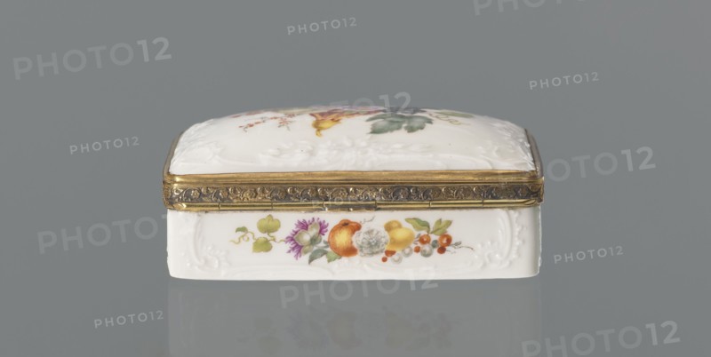 Snuffbox tin, Koenigliche Porzellan Manufaktur KPM; Berlin; 1763–1918. Snuffbox in the form of a rectangular box with a slightly arched lid, framed in gilded bronze, with a plant ornament around the outer edge of the lid and a lock, on hinges. Around the edge of the lid and the side walls of the tin there is a relief with rocaille and floral motifs. On the lid, the bottom of the base and each of the side walls are painted with colorful fruit and floral compositions including blackcurrant, fig, mirabelle plum or redcurrant and plums. Inside the lid is a painted composition of a bouquet of flowers in a cup-shaped brown vase on a stone tabletop with fruit and a sprig of raspberry flowers; on the edge of the tabletop a small bird sparrow? nibbling a leaf, above two butterflies flying. Ca. 1765–1770. Porcelain haberdashery snuffbox. Germany, Berlin. Height 3.2 cm; width 8.3 cm; depth 6 cm., A small rectangular trinket box sits centered against a gray background, with a white porcelain body ...