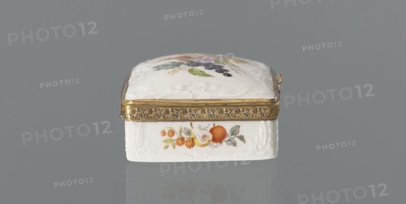 Snuffbox tin, Koenigliche Porzellan Manufaktur KPM; Berlin; 1763–1918. Snuffbox in the form of a rectangular box with a slightly arched lid, framed in gilded bronze, with a plant ornament around the outer edge of the lid and a lock, on hinges. Around the edge of the lid and the side walls of the tin there is a relief with rocaille and floral motifs. On the lid, the bottom of the base and each of the side walls are painted with colorful fruit and floral compositions including blackcurrant, fig, mirabelle plum or redcurrant, and plums. Inside the lid is a painted composition of a bouquet of flowers in a cup-shaped brown vase on a stone tabletop with fruit and a sprig of raspberry flowers; on the edge of the tabletop a small bird sparrow? nibbles a leaf, above two butterflies flying. Ca. 1765–1770. Germany, Berlin. Porcelain haberdashery snuffbox. Height 3.2 cm; width 8.3 cm; depth 6 cm., A small rectangular decorative box sits centered against a gray background, with a white porcelain bo...
