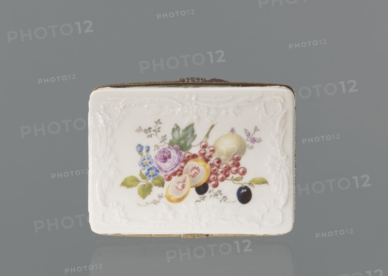 Snuffbox tin, Koenigliche Porzellan Manufaktur KPM; Berlin; 1763–1918. Snuffbox in the form of a rectangular box with a slightly arched lid, framed in gilded bronze, with a plant ornament around the outer edge of the lid and a lock, on hinges. Around the edge of the lid and the side walls of the tin there is a relief with rocaille and floral motifs. On the lid, the bottom of the base and each of the side walls: colorful fruit and floral compositions including blackcurrant, fig, mirabelle plum or redcurrant, and plums. Inside the lid: a colorful composition with a bouquet of flowers in a cup-shaped brown vase on a stone tabletop with fruit and a sprig of raspberry flowers; on the edge of the tabletop a small bird sparrow? nibbling a leaf, above two butterflies flying. Ca. 1765–1770. Porcelain haberdashery snuffbox. Germany; Berlin. Height 3.2 cm, width 8.3 cm, depth 6 cm., A rectangular white porcelain box with embossed decorative borders sits against a gray background, showing a painte...