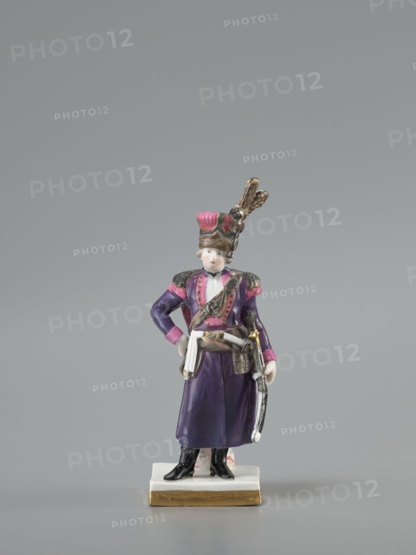 Figurine of an officer of the Polish Guard, Koenigliche Porzellain Fabrique, Meissen 1710–1763, figurine. On a flat square base with gilded side edges, standing leaning against a low marbled column, a man in a purple kontusz with pink lapels, with epaulettes, a sabre strapped to the kontusz belt, a black map case slung on a belt and leaning against the column at the back; on his head is a high pink cap with black decorations and a high broken tail placed at the side. Defined signature on the bottom of the base as a Galician noble guardsman, in the Austrian service. Dated to the 18th/19th century model marking with the letter K 1-100 from ca. 1791. Saxony; Meissen., A small porcelain figurine of a person wearing a tall hat with decorative feathers stands on a rectangular base with a gold-colored edge; the figure wears a purple coat with pink accents on the shoulders and cuffs, a white sash at the waist, black boots, and holds a curved object at the side, with visible colors including pu...