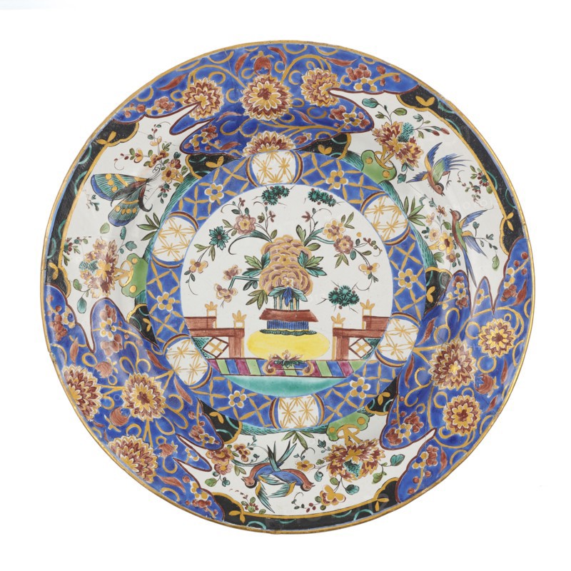 Plate, Karol Wolff faience factory; 1783–1800 maker, Sultan's service imitation, floral decorations, Far Eastern motifs, Stanislaw August Poniatowski King of Poland; 1732–1798, insects. Plate colorfully decorated with Far Eastern floral motifs; imitation of decoration from a Sultan's service produced by the royal factory in Belweder, late 18th century, Bielino, Poland; vessel; height 3.6 cm; width 25.0 cm, A round decorative plate with a central circular scene surrounded by patterned borders, featuring a floral arrangement in a vase at the center with a few stylized flowers and leaves in shades of yellow, pink, green, and blue, set on a base with brown and yellow tones; the inner ring around the central scene shows geometric and floral motifs in blue, gold, and white, and the outer rim is decorated with large floral designs and scrolling shapes in vivid blue, gold, brown, and touches of green and black, along with several small birds and butterflies in blue, green, and pink hues agains...