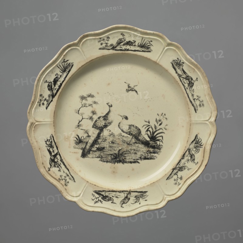 Plate with a pattern of Liverpool birds, Sadler & Green Liverpool, Josiah Wedgwood's Brick House Works, Derby Pot Works, peacocks, Liverpool birds, print, Wedgwood, Josiah 1730–1795. Plate with a 6-part collar with a Catherine relief around the edge and decoration in black print, a pattern of exotic Liverpool birds in the mirror and on six segments of the collar a peacock and pheasant motif in a summed landscape. c. 1765–1770, England, possibly Burslem or Derby. Earthenware plate, height 2.2 cm, diameter 24.0 cm., A scalloped ceramic plate with a pale cream surface and black decorative illustrations, featuring a central scene of two birds standing on a grassy mound surrounded by small plants, a few scattered marks on the surface, and a few smaller bird and foliage motifs arranged around the rim; the background is a solid gray.