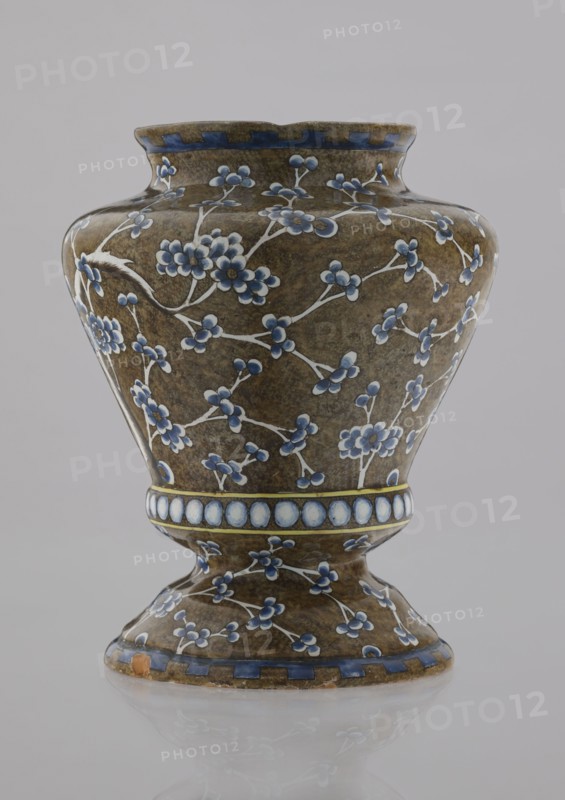 Vase by Karol Wolff fl. ca 1800; Bielino faience factory 1779–1800. A baluster-shaped Warsaw faience vase decorative vessel, profiled at the bottom with a short, slightly flared neck and a vaulted, profiled foot. Olive-brown body with a white, cobalt-shaded depiction of a plum tree whose bent, widely spread branch and network of flowery twigs cover the surface. Around the profiled belly is a row of white, shaded pearls; the rim bears a stylized teeth ornament. Chinoiserie/Far Eastern motifs, plant decorations. Inscribed/dated stylistically 1770–1800. Height 17.0 cm; width 13.2 cm. Origin: Warsaw, Poland., A ceramic vase with an hourglass-like silhouette sits against a pale gray background, its surface covered in a mottled brown glaze decorated with a pattern of white branches and small blue flowers and buds; the rim and base edges have a band of blue rectangles, and a central band around the narrowest part shows a row of pale blue circular medallions outlined in yellow, while the overa...