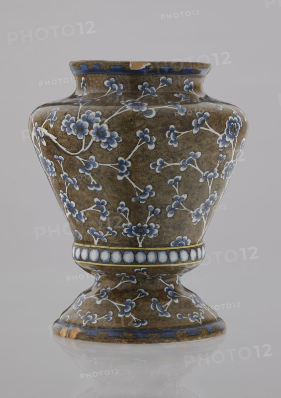 Vase, Karol Wolff fl. ca. 1800 author, Bielino faience factory 1779–1800 author; baluster-shaped vase with profiled bottom, short slightly flared neck, vaulted profiled foot; body olive-brown with white, cobalt-shaded depiction of a plum tree whose bent, widely spread branch covers the surface with a network of flowery twigs; row of white, shaded pearls around the profiled belly; edge ornament of stylized teeth; chinoiserie/Far Eastern motifs; plums; plant decorations; decorative vessel; 1770–1800; Warsaw, Poland; height 17.0 cm, width 13.2 cm, A ceramic vase with a rounded body and flared neck sitting on a pedestal base, covered in a brown mottled glaze and decorated with a pattern of small floral sprigs in shades of blue and white with pale stems, a band of circular motifs in white and blue encircling the midsection, and blue geometric border accents at the rim and base, with a small chip on the rim and another on the base, photographed against a plain light gray background.