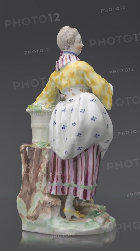 Figurine: gardener, Kaiserliche Porzellanmanufaktur in Wien 1744–1864 manufacturer, On an irregular, rocky base, a young woman in a gardener's outfit leans her left arm on a tall basket of flowers standing on a rocky base or a tree trunk?, holding twigs with leaves in her hands; leaning slightly to the left, with her left leg bent at the knee, resting on a rocky ledge. Dressed in a beetroot-striped skirt trimmed with green ribbon at the bottom, a tied-up apron with blue flowers, a short yellow jacket with flared sleeves, from under which the puffs of a white blouse peek out, a bodice with a gilded pattern, a cherry-striped scarf tied in a knot at the neck, with long twisted ends falling to the waist and a blue ribbon with a bow at the neck; hair pinned up and combed smoothly. ca. 1770, Vienna, Austria; porcelain figurine, height 20 cm, A porcelain figure of a woman standing beside a short tree stump, shown from the back and slightly in profile, wearing a yellow jacket with a marbled ap...