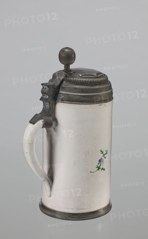 Cylindrical tankard in a pewter frame, Prószków Slaski faience factory 1763–1853. White, painted in colors. From the front, a depiction of a barefoot woman sitting under a tree, her blouse unbuttoned, revealing her breasts. The woman, head turned to the right, supports with her right hand a small child, dressed in a short T-shirt, running towards her from the left. It can be interpreted as an allegorical representation of Maternitas motherhood. The representation is painted with some freedom. Late 18th century, Silesia Province of Silesia, 1742–1918. Cylindrical tankard, vessel. Height 24.0 cm, width 15.0 cm, depth 10.5 cm., A cylindrical ceramic tankard with a pale off-white surface and a curved handle on the left is shown against a neutral gray background; it has a metal hinged lid topped with a round metal knob and decorative ridged banding, with matching metal fittings at the handle attachment and base, and a small painted floral motif in blue and green on the body.