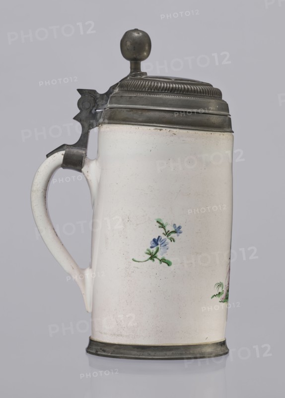 Cylindrical tankard in a pewter frame. White faience painted in colors. From the front, a barefoot woman sitting under a tree, her blouse unbuttoned, revealing her breasts; head turned to the right, she supports with her right hand a small child, dressed in a short T-shirt, running towards her from the left—interpreted as an allegorical representation of Maternitas motherhood. Painted with some freedom. Late 18th century, Prószków Slaski faience factory; 1763–1853, Province of Silesia 1742–1918, Silesia. Vessel. Height 24.0 cm; width 15.0 cm; depth 10.5 cm., A ceramic tankard with a hinged metal lid and thumb lever, shown in profile against a neutral background; the body is off-white with a glossy surface and small scattered imperfections, featuring a small floral painted motif in blue and green on its side and a hint of another motif at the edge, a curved off-white handle attached to the side, and metal bands at the base and around the top with a rounded metal finial on the lid; visib...