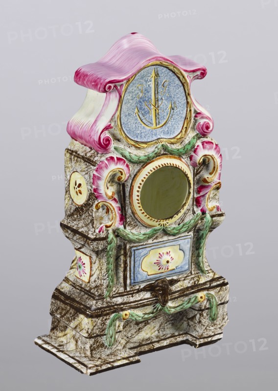 Clock setting, Prószków Slaski faience factory; 1763–1853. Clock setting in the shape of a late Baroque stove supported by a low base. Marbled, decoratively decorated, three-story, supported by a low base. In the central storey, above the pedestal with doors, is a circular recess for a watch framed by rocailles and laurel garlands. The volute-topped section features a shield with the monastic inscription JG/W against an anchor in the background. Painted colorfully in brown and purple tones, with yellow, green and blue accents. Late 18th century, Province of Silesia 1742–1918, Silesia, Prószków Slaski. Clock setting; height 25.0 cm, width 15.1 cm, depth 5.0 cm., An ornate decorative object with a sculpted form and a curved top painted pink and white, featuring a round central opening surrounded by gold and a blue circular panel above with a gold anchor-like motif, flanked by pink shell-like scrolls and green leafy garlands; the body has a marbled stone-like texture in beige and brown wi...