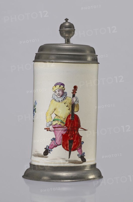Tin-mounted tankard, Prószków Slaski faience factory 1763–1853. White cylindrical tankard in a pewter mount. On the front is a depiction of an obese musician playing a double bass, shown in a vigorous movement. The musician wears a yellow jacket, purple pantaloons, and a lilac hat with a yellow brim. Floral sprigs on the sides. On the tin cover engraved CGH/1811. Late 18th century. Province of Silesia 1742–1918. Dimensions: height 24.0 cm, width 15.0 cm, depth 9.5 cm., A tall cylindrical ceramic stein with a metal lid and base is shown against a plain light gray background; the ceramic body is off-white and features a painted figure of a person wearing a yellow top, purple hat, pink pants, blue bows on dark shoes, and holding a large red stringed instrument with a brown neck and black endpin, with a few small blue and green floral accents visible at the side.