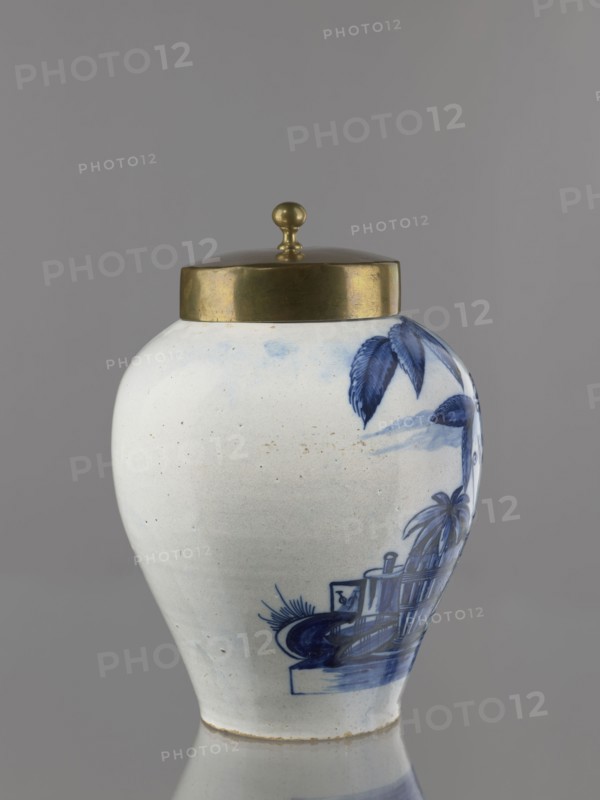 Tobacco jar with brass lid. Ovoid faience vase with a low, slightly parted neck, painted in cobalt on white. Genre scene depicting an Indian smoking a pipe under a tobacco bush and a vase labeled No. 17, arranged against a schematic seascape with two sailing ships. An Indian sits on a pedestal on which stands the labeled ovoid vase with lid; on the opposite side of the pedestal is a basket with a pineapple and a container. Similar vessels serving as tobacco jars were produced in the Netherlands throughout the 18th century. Inscriptions on the vase on the pedestal likely refer to the origin or type of tobacco. 18th century, Netherlands, Delft. Vessel: height 25 cm, width 21.5 cm; lid: height 7.6 cm, width 12.8 cm., A rounded ceramic jar with a domed metal lid topped by a small spherical finial sits against a plain gray background, the jar showing a white base color with blue painted decorative motifs including stylized leaves and a scene with architectural and plant forms, and the metal...