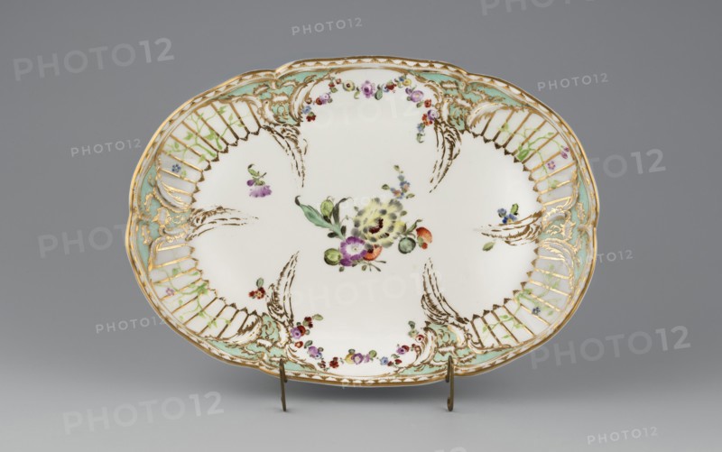Vase base with Reliefzierat decoration; manufacturer: Königliche Porzellan-Manufaktur KPM, Berlin; ca. 1770–1780. Oval, bowl-shaped base with a relief of six triangular flame rocailles with celadon centers around the inner edge, separating four fields featuring a trellis of gilded stamens with an interwoven green branch with small flowers Reliefzierat. Two fields on the longer sides contain colorful floral garlands; the center mirror has a loose floral arrangement. Adjacent to the rocailles are scattered small flower sprigs. Around the edge of the base is a gilt strip separated by a half-cylinder. Maker association: Frederick II Frederick the Great, King of Prussia, 1712–1786. Origin: Berlin, Germany. Function: vessel., An oval ceramic platter sits on a small metal stand against a neutral gray background, primarily white with decorative painted floral motifs in the center and around the rim. The rim features gilded scrollwork and lattice-like cutout patterns accented with pale green an...