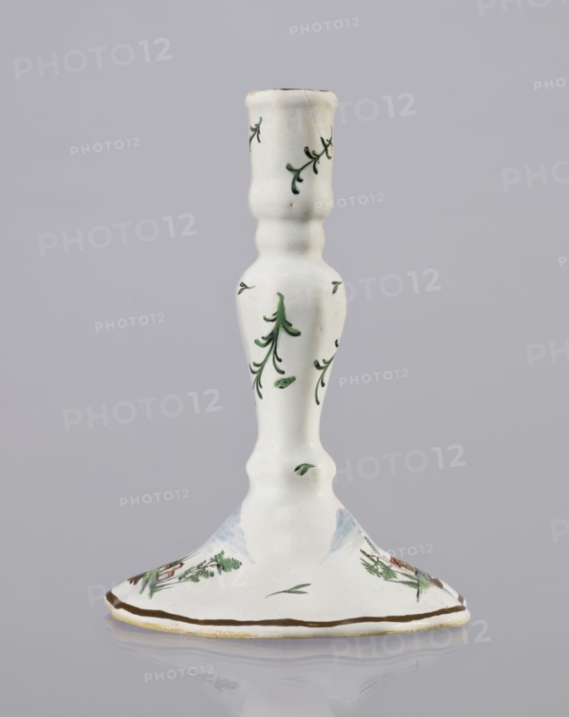 Candlestick, Prószków Slaski faience factory active 1763–1853. Candlestick in the shape of a polygonal baluster supported on a wide, round conical base with a wavy edge. Painted colorfully; on the foot are two miniatures depicting landscapes with architecture and oblique projections of twigs on the shaft. Late 18th century, circa 1770–1783. Silesia Province of Silesia. Prószków Slaski. Candlestick; height 18.2 cm, width 13.0 cm., A white ceramic candlestick with a tall, slender, turned shape and a flared base, decorated with small green leafy sprigs and simple painted foliage; the base shows additional hand-painted green and brown landscape-like motifs and a thin brown rim, and the overall colors visible are white, green, brown, and a touch of pale blue.