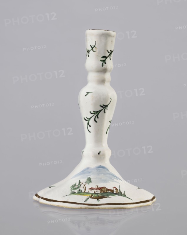 Candlestick, Prószków Slaski faience factory 1763–1853. Candlestick in the shape of a polygonal baluster supported on a wide, round conical base with a wavy edge. Painted colorfully; on the foot are two miniatures depicting landscapes with architecture, and oblique projections of twigs on the shaft. 1770–1783. Silesia Province of Silesia. Prószków Slaski. Porcelain/faience candlestick, height 18.2 cm, width 13.0 cm., A white ceramic candlestick with a rounded base and a slender, contoured stem decorated with small green leafy sprigs; the base shows a painted landscape scene with a small house, trees, and distant hills in muted greens, browns, blues, and a touch of red, and a thin brown rim around the edge.