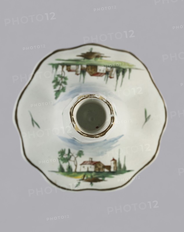 Candlestick, Prószków Slaski faience factory 1763–1853. Candlestick in the shape of a polygonal baluster supported on a wide, round conical base with a wavy edge. Painted in color. On the foot are two miniatures with landscapes featuring architecture; oblique projections of twigs on the shaft. Circa 1770–1783. Prószków Slaski, Silesia Province of Silesia. Height 18.2 cm; width 13.0 cm., A top-down view of a white ceramic cup and saucer set with a scalloped rim and hand-painted landscape scenes. The saucer shows a painted cottage scene with trees and a small building in green, brown, red, and blue hues, mirrored on opposite sides; there are a few green brushstrokes and a thin brownish-gold line around the scalloped edge. The cup sits centered on the saucer, showing its circular interior with a worn brownish edge and a muted pale green inside. The background is a flat light gray.