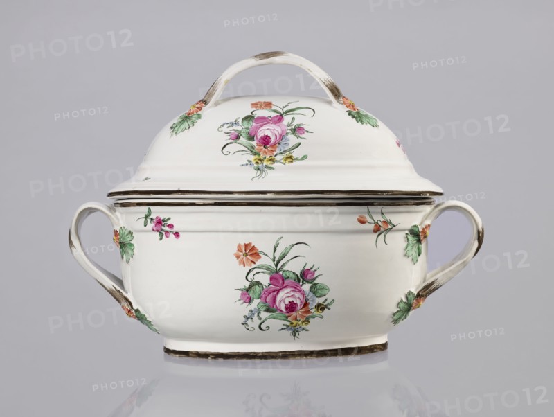 Vase with lid, Prószków Slaski faience factory 1763–1853. Egg-shaped soup tureen with a lid in the form of a flattened ellipsoid/hemisphere. Handles of the vase and lid are in the form of two crossed ribbons. Decorated with small painted floral projections and large bouquets of multicolored flowers. On both sides of the vase and lid are bouquets of flowers with a pink rose. Handles adorned with vermilion daisies with large green leaves. Rim of the vase and cover painted brown. Ca. 1785. Province of Silesia Silesia, Prószków Slaski. Vessel. Height 25.5 cm; length 35.5 cm., A white ceramic lidded tureen with a curved handle on top and two looped side handles, all showing subtle brown brush accents, decorated with floral motifs in pink, green, orange, yellow, and touches of blue, sitting on a reflective surface against a pale gray background.