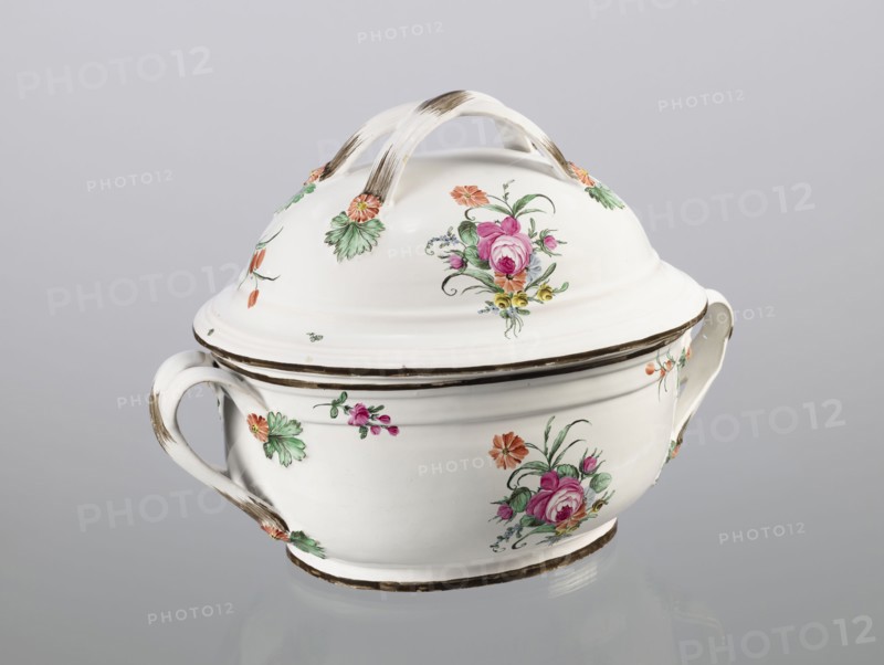 Vase with lid, Prószków Slaski faience factory 1763–1853; egg-shaped soup tureen with lid in the form of a flattened ellipsoid/hemisphere. Handles of the vase and lid are in the form of two crossed ribbons. Decorated with small painted floral projections and large bouquets of multicolored flowers; on both sides of the vase and lid are bouquets with a pink rose. Handles adorned with vermilion daisies with large green leaves. Rim of the vase and cover painted brown. Floral decorations, roses. Circa 1785. Silesia Province of Silesia. Vessel. Height 25.5 cm; length 35.5 cm., A white ceramic covered bowl with a lid and two handles sits against a pale gray background, decorated with painted floral motifs in pink, green, orange, yellow, and a bit of blue, with brownish brush accents on the handles, rim, and base; the lid has a curved double-arched handle and matching floral decorations, and the bowl shows several clusters of flowers and leaves around its sides.