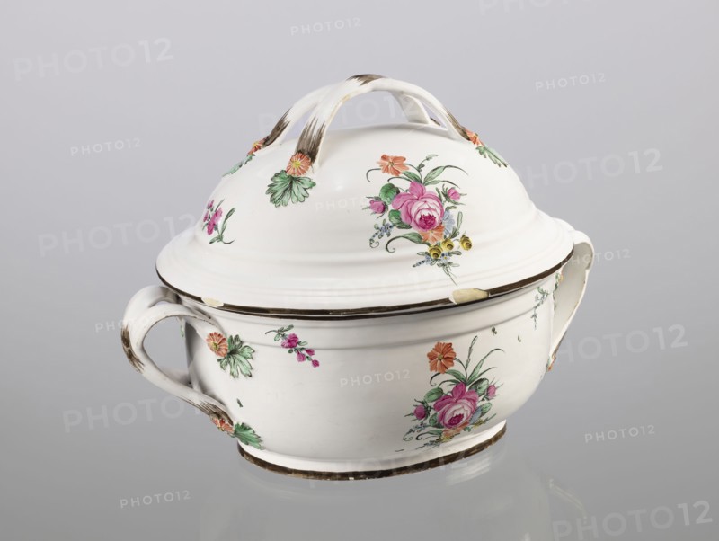 Vase with lid, Prószków Slaski faience factory 1763–1853, egg-shaped soup tureen with a lid in the form of a flattened ellipsoid/hemisphere. Handles of the vase and lid are two crossed ribbons. Decorated with small painted floral projections and large bouquets of multicolored flowers; on both sides of the vase and lid are bouquets with a pink rose. Handles are adorned with vermilion daisies with large green leaves. Rim of the vase and cover painted brown. Ca. 1785, Province of Silesia Silesia, Prószków Slaski. Vessel; height 25.5 cm, length 35.5 cm., A lidded ceramic tureen with a glossy white surface sits against a pale gray background, featuring a curved handle on each side and an arched double handle on the domed lid; the piece is decorated with painted floral motifs in pink, green, orange, yellow, and touches of blue, and shows areas of brownish discoloration or wear along the handles and rims.
