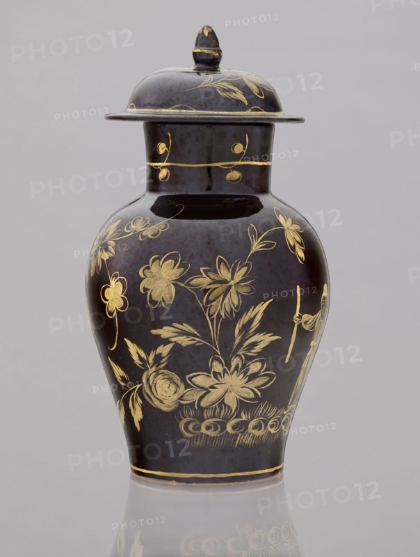 Kuan-type vase with cover, Bielino faience manufactory; 1779–1800. Maker: Karol Wolff fl. ca. 1800. Chinoiserie, Chinese/Far Eastern motifs, Warsaw faience, unknown provenance. Low kuan-type vase in the shape of a bulging baluster with a cylindrical neck and a low domed cover with an acorn-shaped handle. Against a chocolate background a gold-painted scene: a fragment of a garden with two chrysanthemum bushes, a peony bush, a palm tree, a rice fence, and between them the figure of a Chinese man with a bag and a walking stick. Around the neck is a thin simple plant twig; a painted flowered branch on the lid. Late 18th century, Poland. Vessel: height 19.5 cm, diameter 13.5 cm; lid: height 7.5 cm, diameter 10.5 cm., A lidded vase with a glossy dark surface decorated with floral and botanical designs in metallic gold tones; the vase has a rounded body that tapers toward the base and neck, a flared rim under the lid, and a domed lid with a small knob on top also decorated in gold; the visibl...