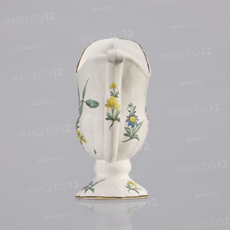 Nalewka, Prószków Slaski faience factory; 1763–1853, maker/manufacturer markings; floral decorations, locking, carnations, helmet-shaped nalewka with locking and fluted surface. Bowl base and vaulted foot modeled with four leaves. T-shaped handle. Colorful decoration: bouquet with a large purple carnation with three buds and three small yellow flowers; small projections of flowers and leaves. Brown borders along the handle, edge of the bowl, and foot. Dated 1770–1783. Region: Silesia Province of Silesia. Prószków Slaski. Vessel; height 19.0 cm, width 19.0 cm, depth 9.5 cm., A white ceramic pitcher with a handle and a short pedestal base is shown against a light gray background. The pitcher has hand-painted floral decorations in yellow, blue, green, and a touch of purple, with green stems and leaves. The rim and base edge have a thin gold trim. The pitcher’s surface shows subtle glaze variations and a glossy finish, and there is a soft reflection of the base on the surface beneath.