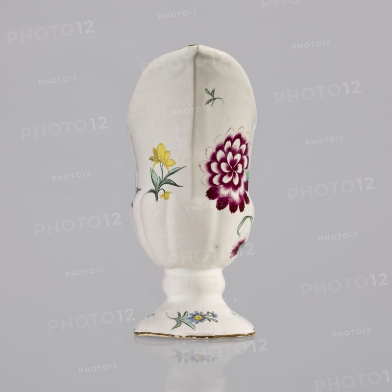 Nalewka, Prószków Slaski faience factory; 1763–1853, maker/manufacturer; floral decorations; locking; carnations flowers; repossession; Helmet-shaped Nalewka with locking and fluted surface. Bowl base and vaulted foot modeled with 4 leaves. T-shaped handle. Colorful decoration — bouquet with a large purple carnation with 3 buds and 3 small yellow flowers; small projections of flowers and leaves. Brown borders along the handle, edge of the bowl, and foot. Dated 1770–1783. Region: Province of Silesia, Silesia, Prószków Slaski. Vessel; height 19.0 cm, width 19.0 cm, depth 9.5 cm., A white ceramic vase with a scalloped elongated body and a rounded pedestal base, decorated with painted floral motifs in yellow, green, blue, and deep magenta, showing a large magenta flower on one side and smaller yellow and blue flowers with green leaves around the body and base, set against a neutral pale gray background.