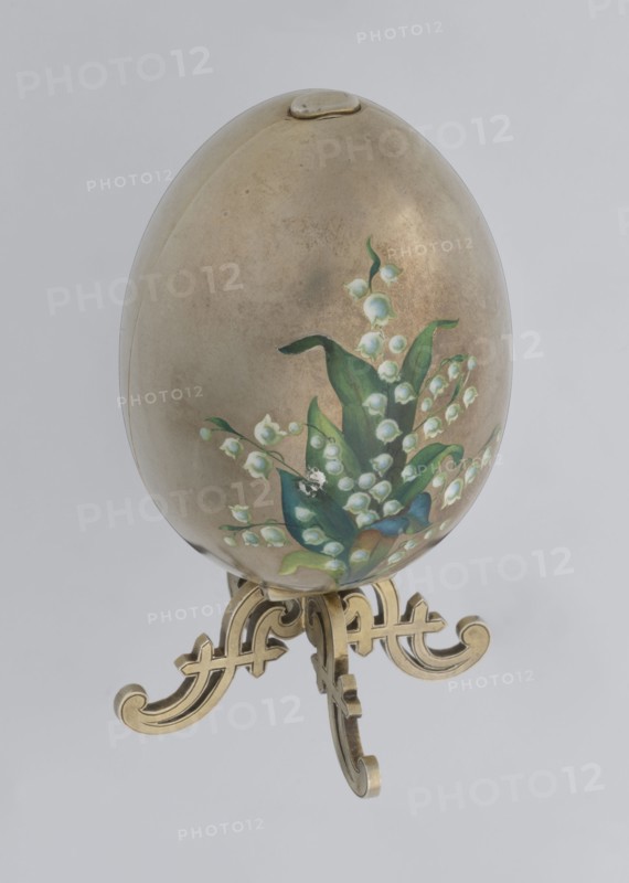 Easter egg, silver, smooth, composed of two opening halves on a hinge, cut vertically; on one part an enameled bouquet of forget-me-nots; gilded interior, with an octagonal basket for embedding a surprise missing. Egg supported on a four-legged stand. Decorated with lilies of the valley and flowers., Date: 1890s, St. Petersburg. Decorative object. Height 11.4 cm, width 6.6 cm, depth 6.6 cm, weight 233.5 g., An egg-shaped object with a metallic beige surface sits on an ornate stand; the egg features a painted floral cluster of bell-shaped blossoms and broad leaves in shades of green, white, and touches of blue, with the stand rendered in a gold color and the background a pale gray.
