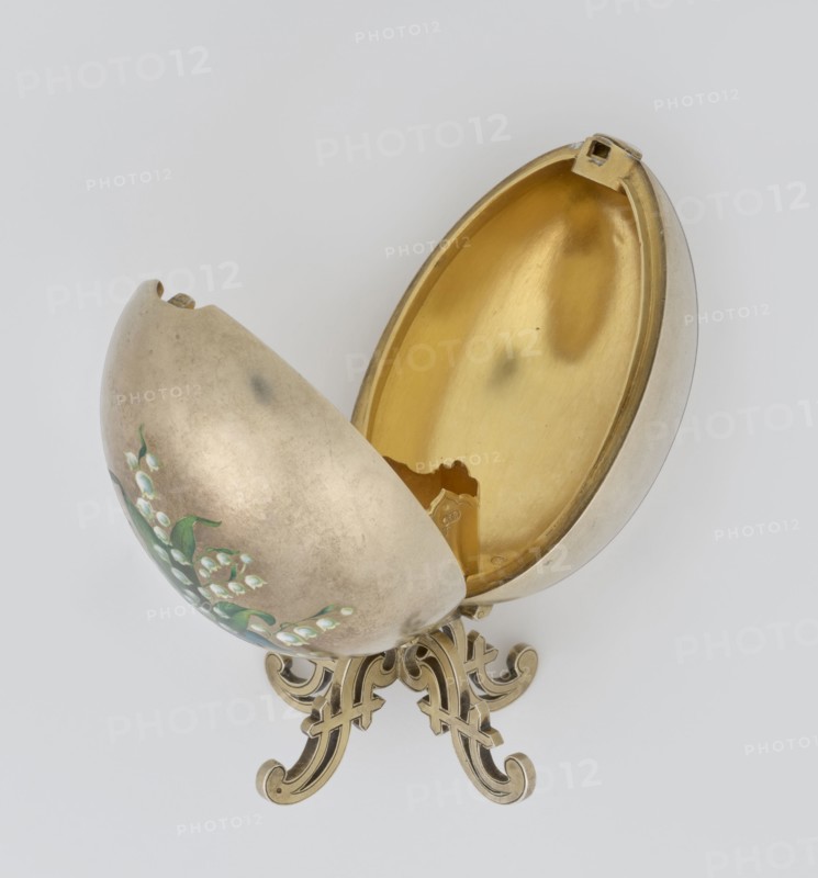 Easter egg, lilies of the valley and flowers motif; silver egg, smooth, composed of two opening halves on a hinge, cut vertically; on one part an enameled bouquet of forget-me-nots; gilded interior with an octagonal basket for embedding a missing surprise. Egg supported on a four-legged stand; 1890s, St. Petersburg; decorative object. Height 11.4 cm, width 6.6 cm, depth 6.6 cm, weight 233.5 g., A decorative egg-shaped object sits open on an ornate stand, showing a shiny gold-colored interior and a muted silver-gray exterior with painted green leaves and small white bell-shaped flowers; the stand and inner rim have warm golden tones, and the background is a pale light gray.