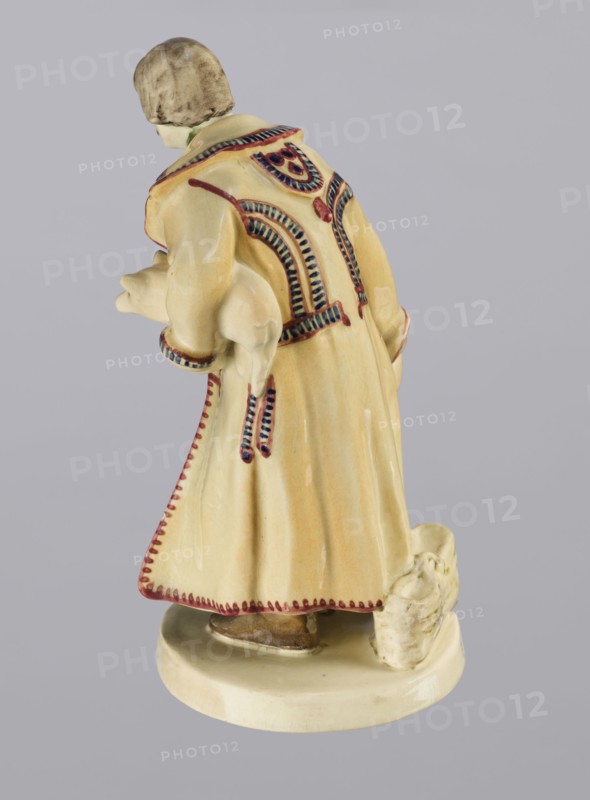 Peasant with Piglet, First Galician Factory of Artistic Faience and Terracotta Pacyków; 1912–1939 manufacturer, Popiel, Antoni 1865–1910 sculptor. Figurine of a man in a peasant Hutsul russet coat on a circular base. The peasant, with straight-cut hair and a period mustache, stands slightly bowed, holding a hat in his right hand and a piglet under his left arm. A woven basket is on the base at the back. Warm tones: light ochre russet coat with brown-purple trim; underneath a green vest, dark trousers, and brown shoes. ca. 1910. Pacyków. Terracotta figurine. Height 14.5 cm; width 8.0 cm; depth 5.5 cm., A ceramic figurine viewed from behind showing a person wearing a long coat with decorative trim, holding a small animal under one arm and standing on an oval base; the coat is primarily pale yellow with red and dark blue decorative accents, the small animal and base are off-white, the person's hair is a muted brown, and there are subtle brown and beige tones on the figure and base.