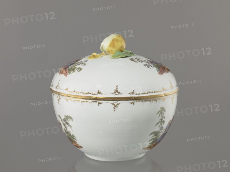 Punch vase with lid, Königliche Porzellan-Manufaktur KPM, Berlin; 1763–1918 maker marking; porcelain with reliefs and enamel genre miniatures. Hemispherical bowl with bottom rim and arched circular lid topped by a plastic half-peeled lemon with stem, leaf, two slices and rolled-up peel. Rim of vase and lid with gilded relief rocaille decoration. Two color miniatures each on bowl and lid after engravings by W. Hogarth?: on the lid—1 three men in 18th-century costume seated at a table with a vase and glasses, 2 three men at a table with a vase, one standing; on the bowl—1 six people seated at a round table with glasses and pipes, one figure reclining in front of the table, 2 seven people seated at a table singing, smoking pipes and conversing. Late 4th quarter of the 18th century late 18th century, Berlin, Germany. Vase; height 27.5 cm, width 28 cm., A rounded white porcelain lidded container with a glossy finish sits against a gray background; the lid has a sculpted yellow fruit with gr...