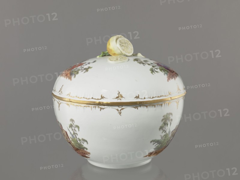 Punch vase with lid, Koenigliche Porzellan Manufaktur KPM; Berlin; 1763–1918. Porcelain, reliefs and colour genre miniatures. Hemispherical bowl with bottom rim, arched circular lid with handle modeled as half a peeled lemon with stem, leaf, two slices and rolled-up peel. Rim of vase and lid with gilded relief rocaille decoration. Two painted miniatures on bowl and two on lid after engravings by W. Hogarth?: lid—park scenes: 1 three men in 18th-century costume seated at a table with a vase and glasses; 2 three men at a table, one standing. Bowl—park scenes: 1 six people at a round table with glasses and pipes, one reclining before the table; 2 seven people at a table, singing, smoking pipes and conversing. 4th quarter of the 18th century, Berlin, Germany. Vase: height 27.5 cm; width 28 cm., A rounded white ceramic covered bowl sits centered against a neutral gray background, the lid topped with sculpted lemon halves and a small green stem in pale yellow and green tones. The bowl and li...