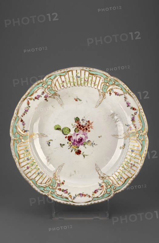 Platter with Reliefzierat decoration and flowers, Königliche Porzellan-Manufaktur KPM, Berlin; maker associated with Frederick II the Great King of Prussia, 1712–1786. Reliefzierat, rocaille ornament; round platter: around the collar, slightly extending into the mirror, a relief of six triangular flamed rocailles with a celadon centre, separating three fields with a trellis of gilded stamens with an interwoven green branch with small flowers Reliefzierat, alternating with three fields with colourful floral decoration and garlands; center of the mirror with a loose floral arrangement, near the rocailles a few small flower sprigs. Half-rolled border with gilding. Ca. 1770–1780, Berlin, Germany. Platter; height 5 cm, depth 34 cm., A round ceramic plate with a scalloped rim displayed on a clear stand against a gray background, the plate having a white base with a central painted bouquet featuring pink, red, and purple flowers with green leaves and small blue accents, the rim decorated with...