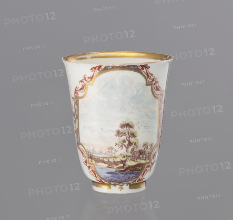 Teapot, Koenigliche Porzellain Fabrique, Meissen c. 1730, 1710–1763, Meissen, Saxony. Teapot, slender, tall, chalice-shaped, without handles, height 7.1 cm, dia. 6.3 cm. Gilded with lace in red and yellow with metallic-violet fields Perlmutterluester. Features a miniature in a Rococo cartouche depicting a riverside landscape; back surface decorated with birds and insects. Maker: Koenigliche Porzellain Fabrique, Meissen., A white porcelain cup with a flared rim and a gold interior edge, decorated with ornate scrolling borders in red and gold and a painted landscape scene on the front; the landscape shows a tree with green foliage, a river or body of blue water, a wooden bridge or fence, grassy banks in shades of green and brown, and a pale sky with soft blue and gray tones; the cup also has gold accents at the base and subtle purple decorative panels along the sides, all set against a neutral gray background.